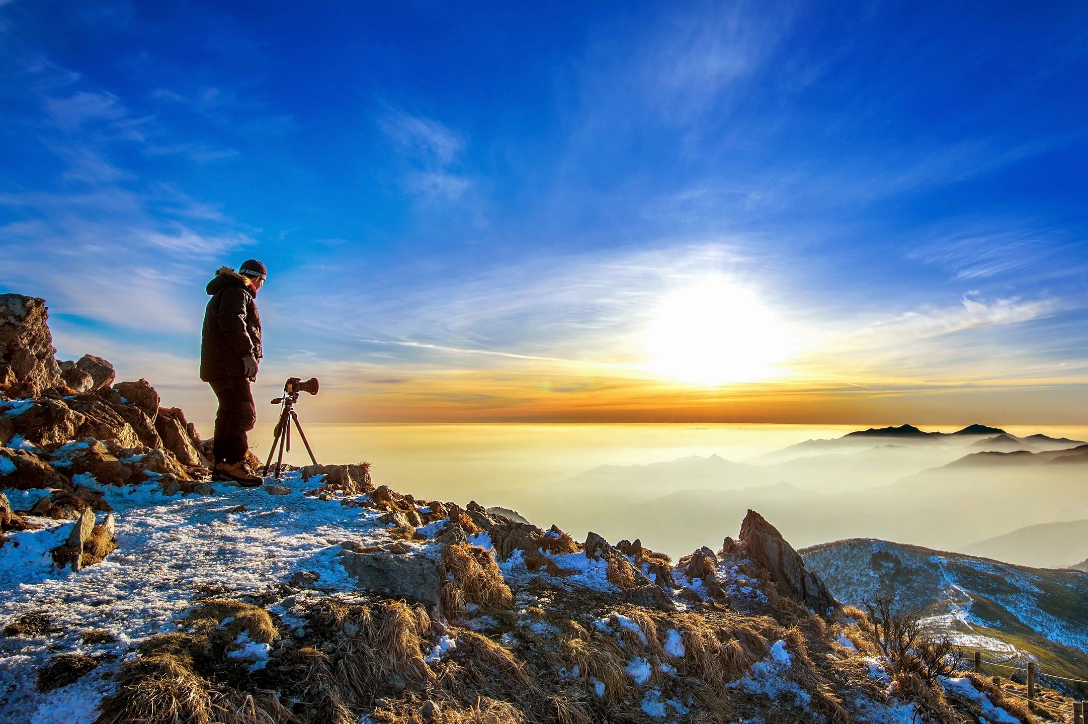 A man is standing on a rock precipice, dressed in a parka, stands behind a camera directed to capture a sunset.