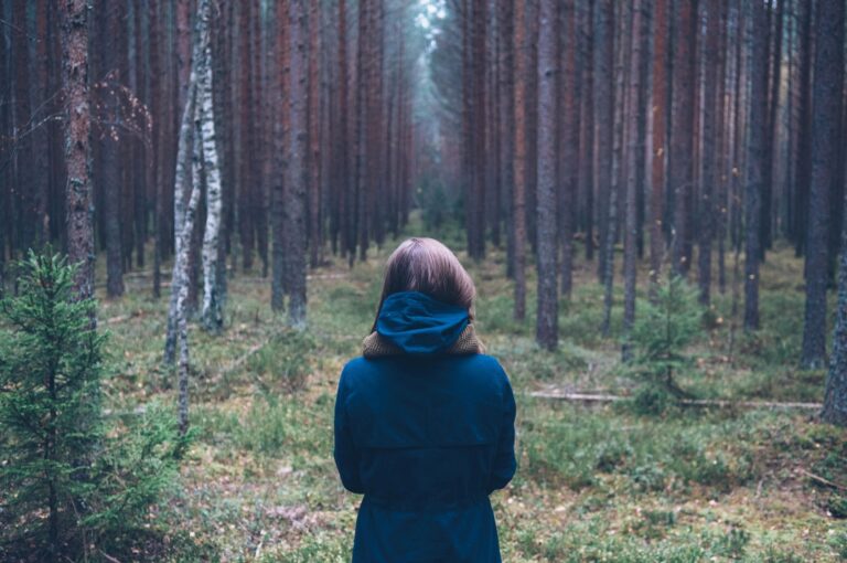 woman stands alone facing a forest of tree trunks